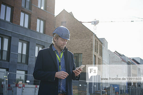Architect wearing hardhat and using mobile phone near buildings
