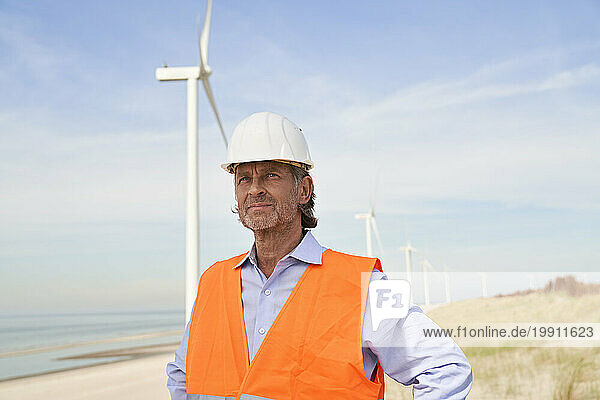 Contemplative engineer wearing hardhat standing in front of wind turbines