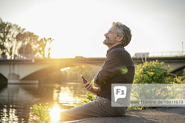 Smiling senior man sitting with mobile phone near lake at sunset