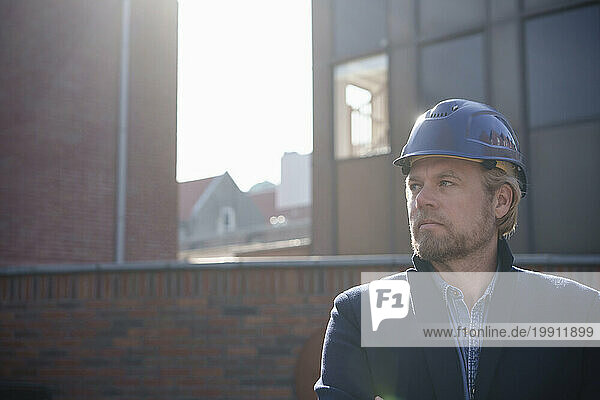 Contemplative architect wearing hardhat in front of buildings on sunny day