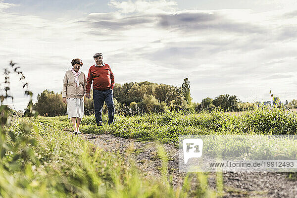 Senior couple holding hands and walking on footpath under cloudy sky