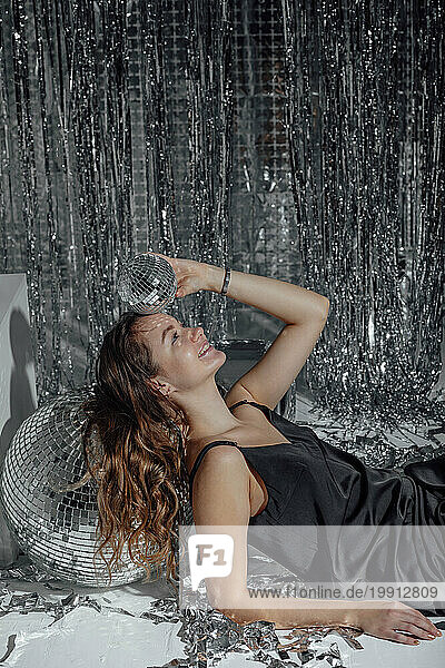 Smiling woman leaning on disco ball near tinsel curtain