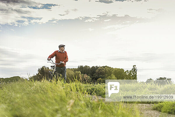 Senior man walking with bicycle on footpath at field under cloudy sky