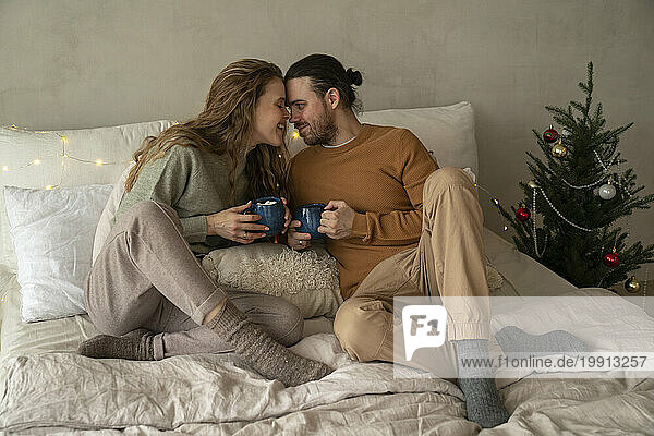 Romantic couple holding mugs on bed near Christmas tree at home