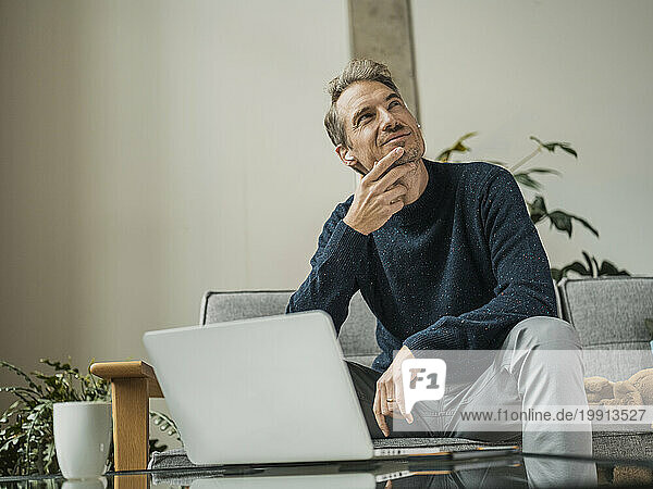 Thoughtful freelancer wearing wireless in-ear headphones sitting near laptop at home