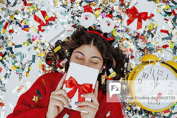 Girl hiding face with gift box and lying on confetti near clock