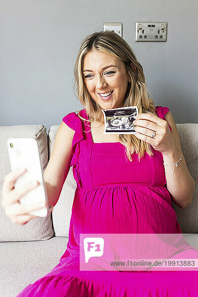 Smiling pregnant woman showing ultrasound photograph on video call through smart phone at home