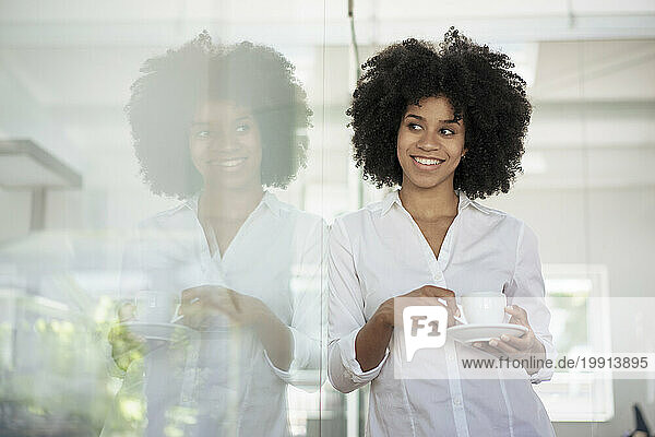 Smiling businesswoman with tea cup leaning on glass wall at office