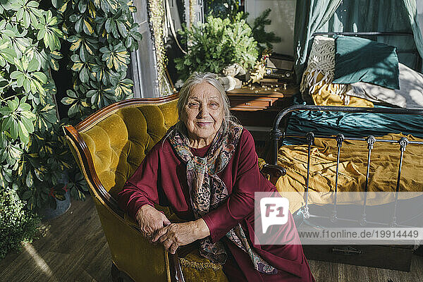 Smiling elderly woman sitting on armchair near bed at home