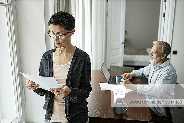 Pensive woman looking at business paper standing next to husband sitting at table working on laptop