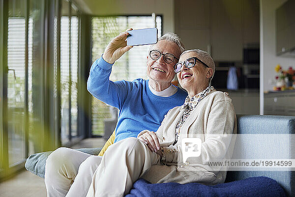 Happy senior couple sitting on couch at home taking selfie
