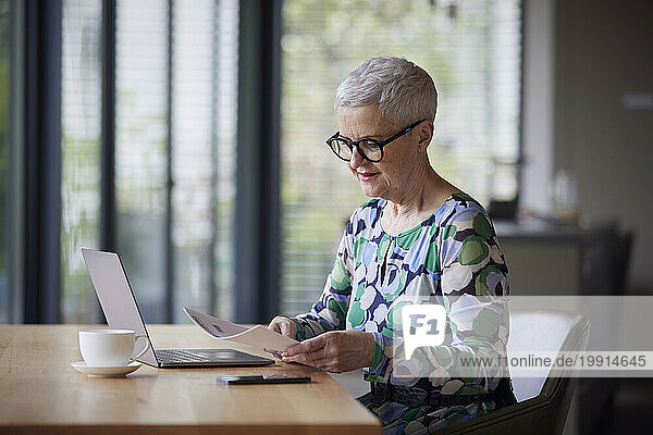 Senior woman with document and laptop at table at home