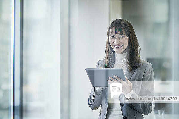 Happy mature businesswoman holding tablet PC seen through glass