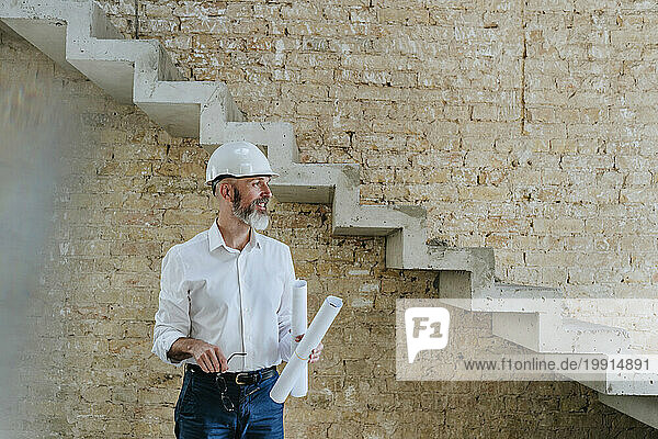 Happy architect wearing hardhat standing with blueprints at site