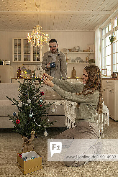 Smiling woman decorating Christmas tree with man standing at home
