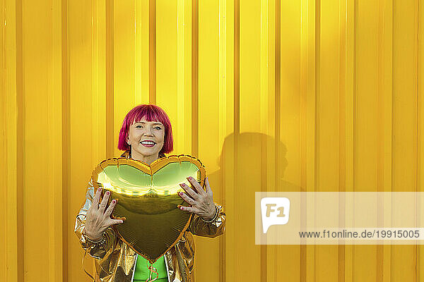 Happy woman holding heart shaped balloon in front of yellow wall