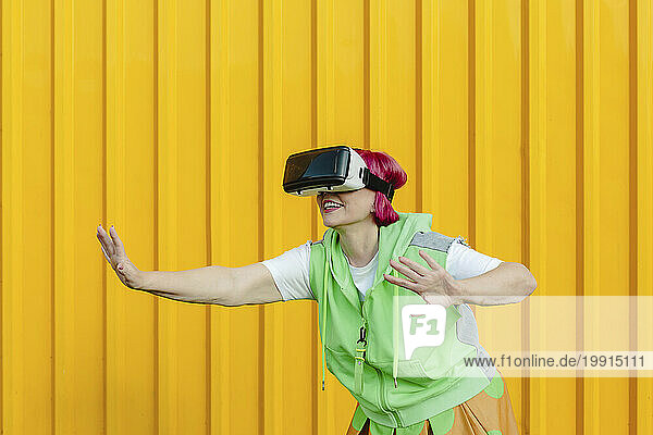 Senior woman wearing virtual reality simulator and gesturing in front of yellow wall