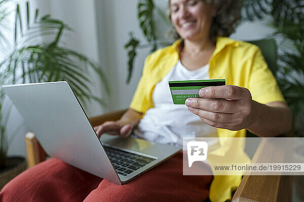 Mature woman holding credit card sitting on armchair using laptop at home