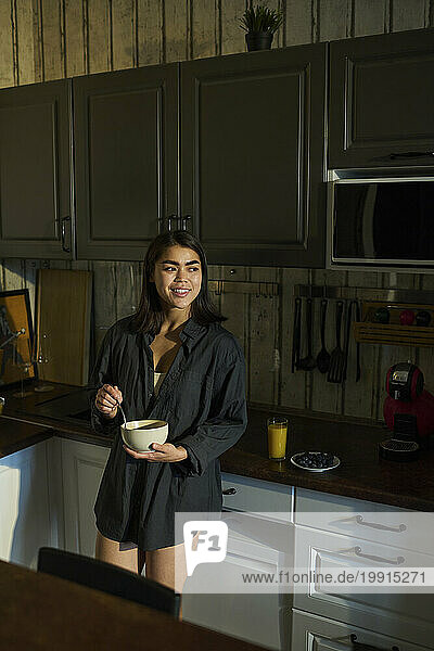 Happy young woman holding bowl of breakfast and standing in kitchen at home