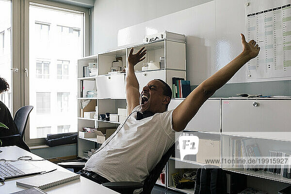 Happy male entrepreneur screaming with arms raised while sitting at office