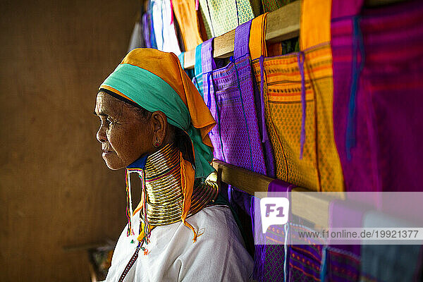 Portrait of senior woman wearing traditional neck rings standing beside colorful clothes hanging on wooden beams  Shan State  Myanmar