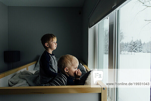 Bright side view of small brothers sitting on bed looking out wi