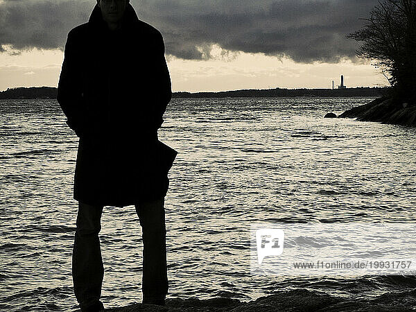A man in a trenchcoat stands at the edge of the water with a factory smokestack in the distance. (silhouette)