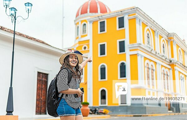 Lächelnde Reisende Frau mit Rucksack  die auf eine Kathedrale auf einem Touristenplatz zeigt. Glückliches Touristenmädchen erkundet die Stadt Granada  Nicaragua  Mittelamerika