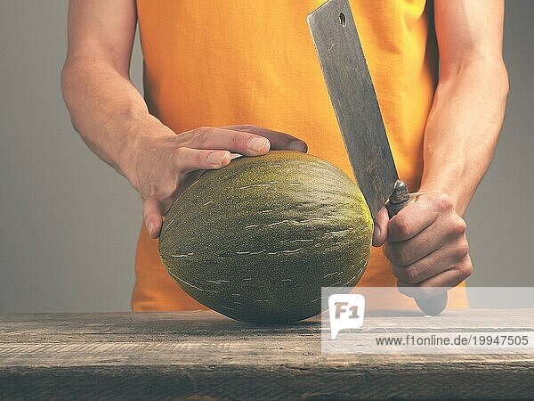 Junger Mann mit einem alten Hackbeil und einer frischen Melone auf einem rustikalen Küchentisch