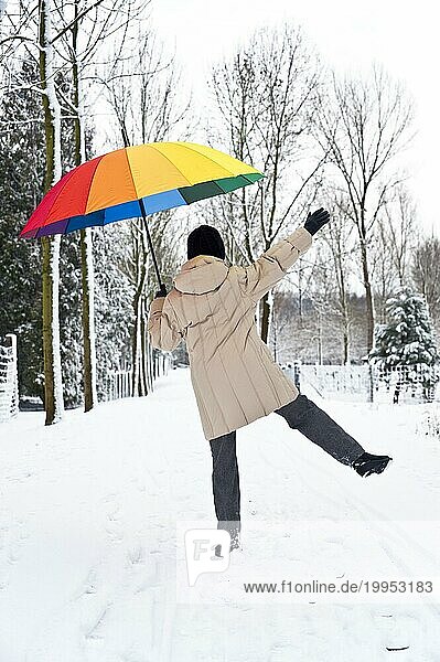 Frau mit buntem Regenschirm springt über einen verschneiten Waldweg