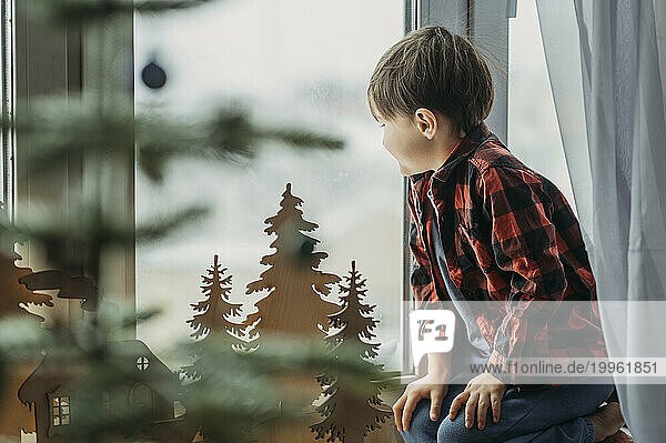 Boy near Christmas decoration looking through window at home