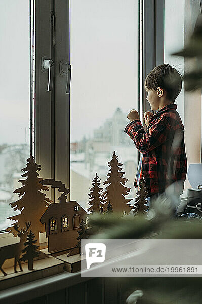 Boy standing near Christmas decoration and looking through window at home