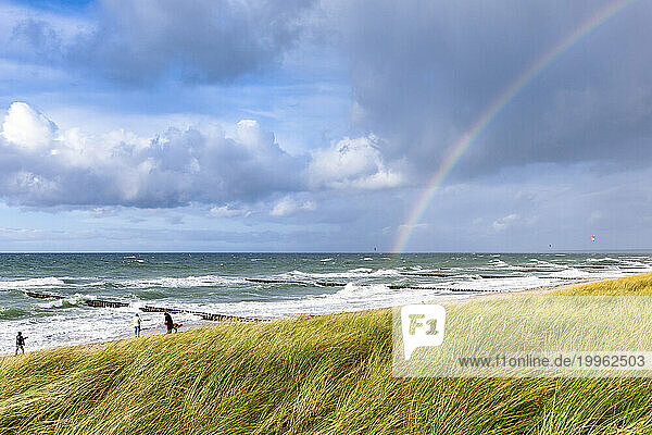 Germany  Mecklenburg-Vorpommern  Grassy beach with rainbow arching against cloudy sky over sea in background