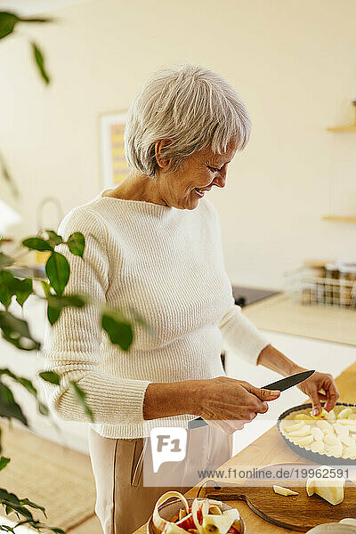 Smiling mature woman cutting apple for pie in kitchen