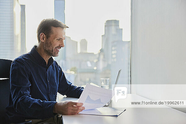 Happy mature businessman sitting with documents and using laptop at desk