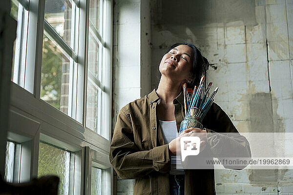 Smiling painter holding paintbrushes with eyes closed standing by window at workshop