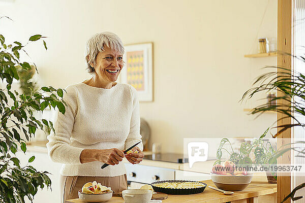 Happy mature woman cutting apple for pie in kitchen