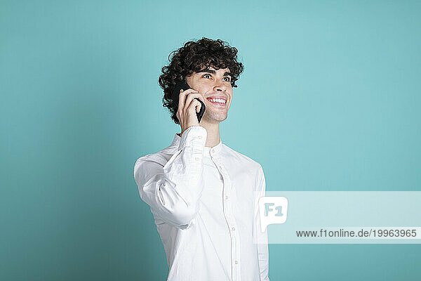 Smiling man talking on smart phone and day dreaming against cyan background