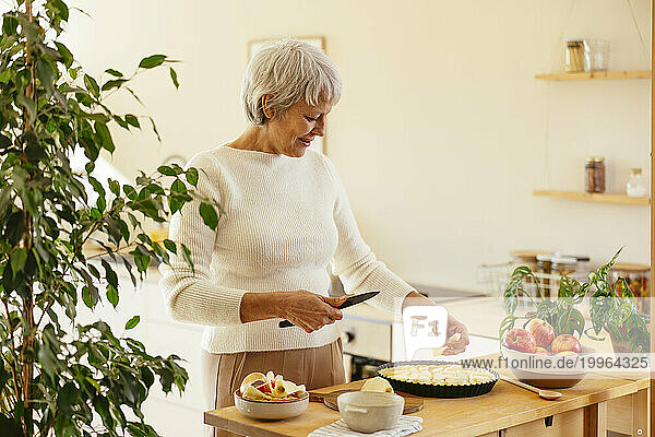 Smiling mature woman cutting apples for pie in kitchen at home