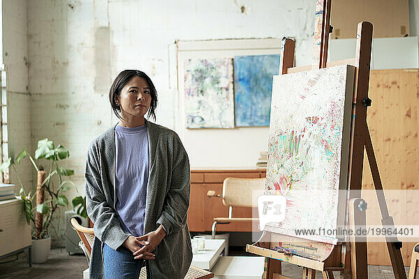 Confident artist standing by painting on easel in workshop