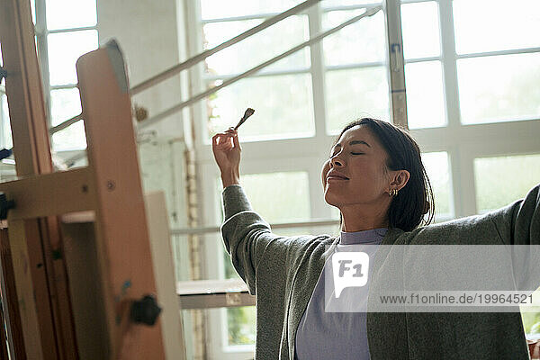 Smiling woman with eyes closed and arms outstretched standing in front of painting at workshop