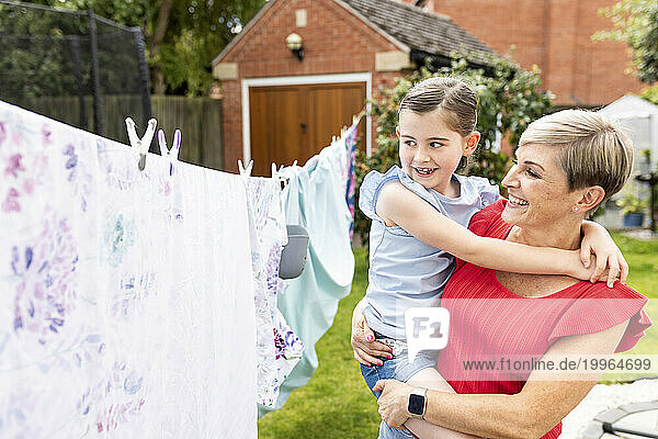 Happy woman carrying daughter near clothes hanging on clothesline in backyard