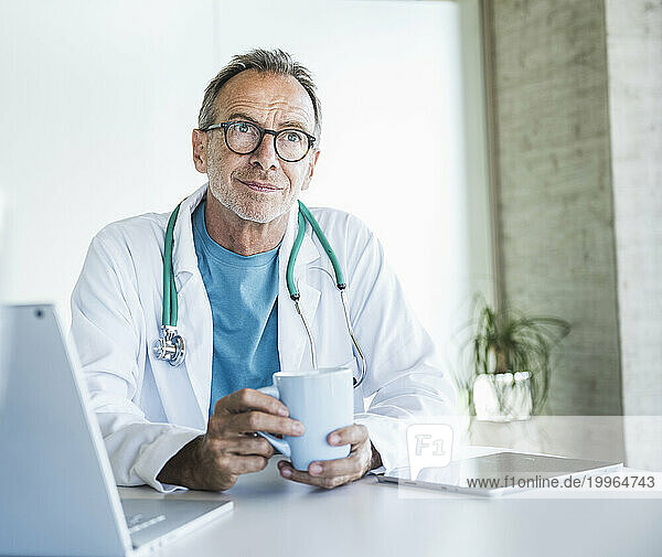 Thoughtful senior doctor sitting with coffee cup at desk