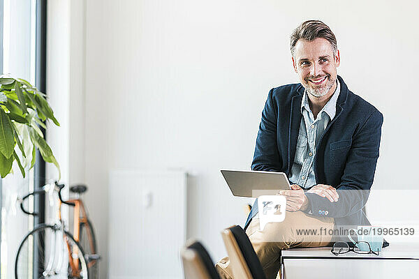 Smiling businessman holding tablet PC and sitting on desk in office