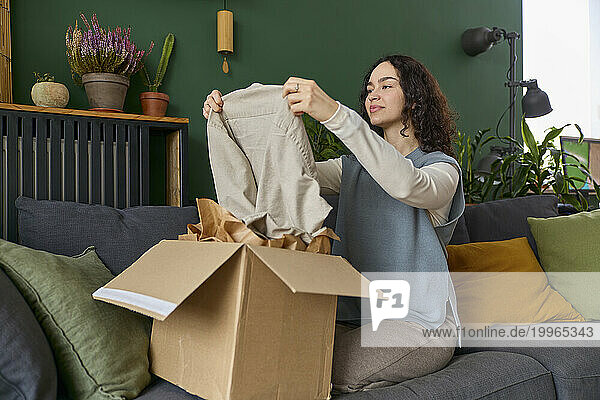 Woman unpacking dress from box sitting on sofa at home