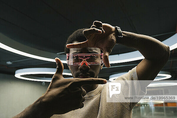Young man with futuristic cyber glasses under modern ring lamp looking through finger frame