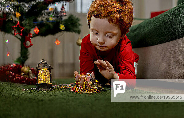 Cute redhead boy playing with Christmas ornaments on green carpet at home