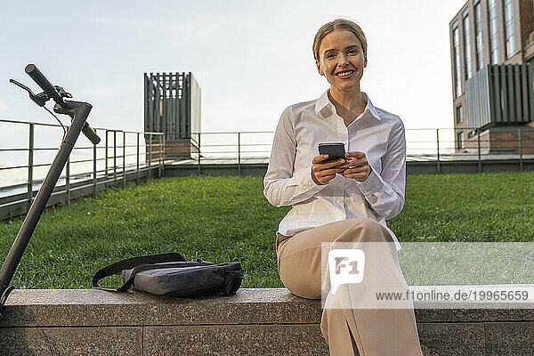 Smiling businesswoman with mobile phone sitting on wall