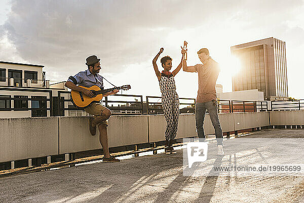 Happy man playing guitar with friends dancing on rooftop
