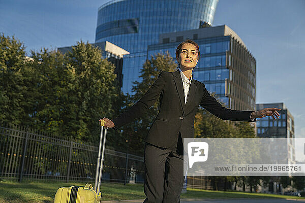 Smiling businesswoman standing with suitcase and hailing ride in city